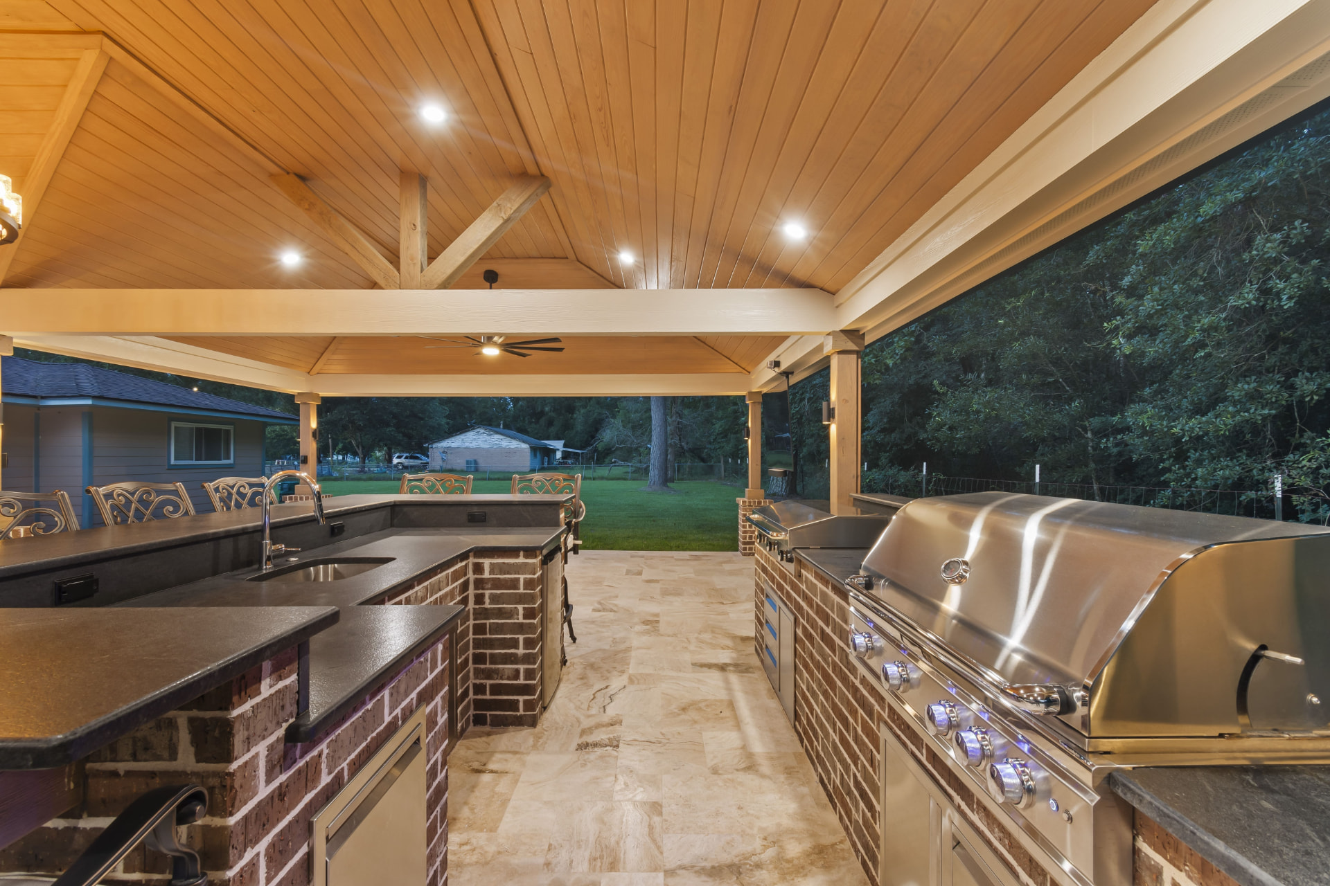 Covered outdoor kitchen with brick base, dark granite counters and stainless grill under vaulted cedar ceiling