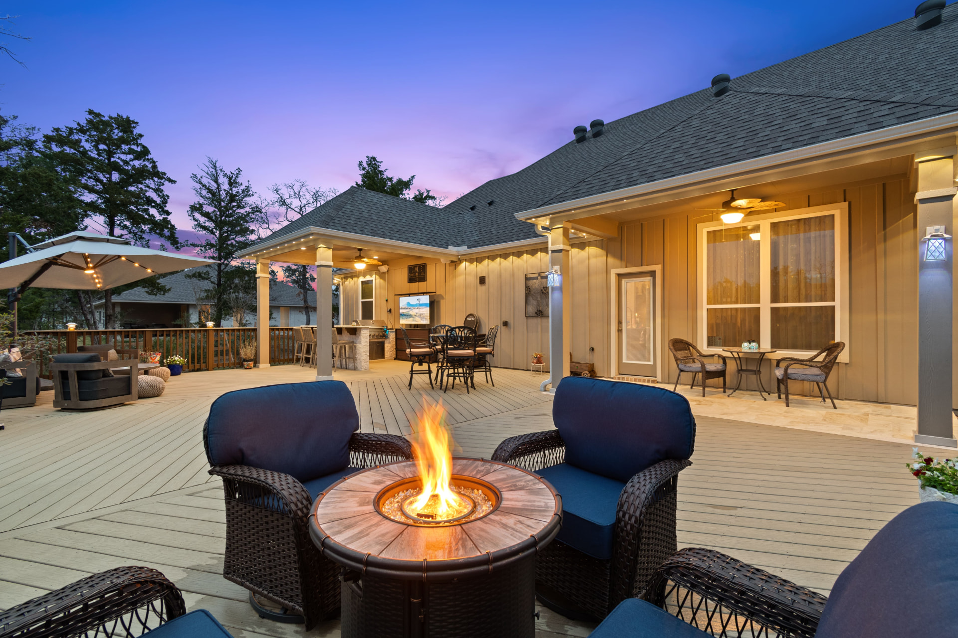 Composite deck at sunset with copper fire table, navy wicker lounge seating and covered outdoor kitchen beyond