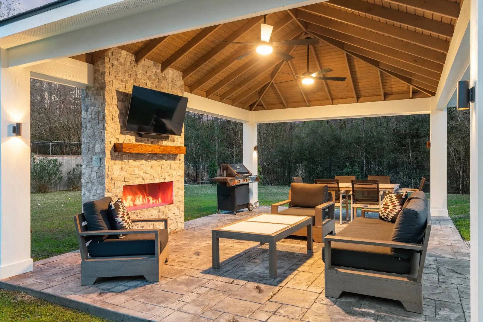 Twilight view of freestanding pavilion with lit stone fireplace and outdoor kitchen