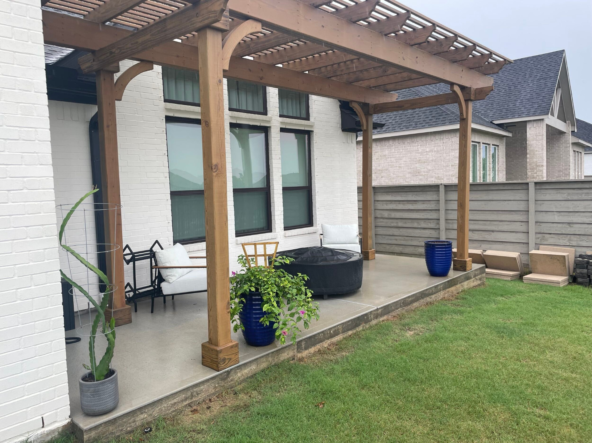 Stained wooden pergola attached to white brick home over concrete patio with seating