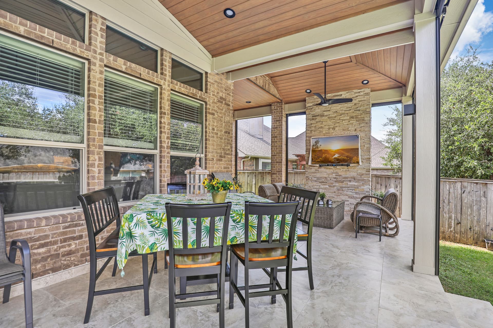 Covered patio with stained wood ceiling, stone fireplace wall with TV, and dining area