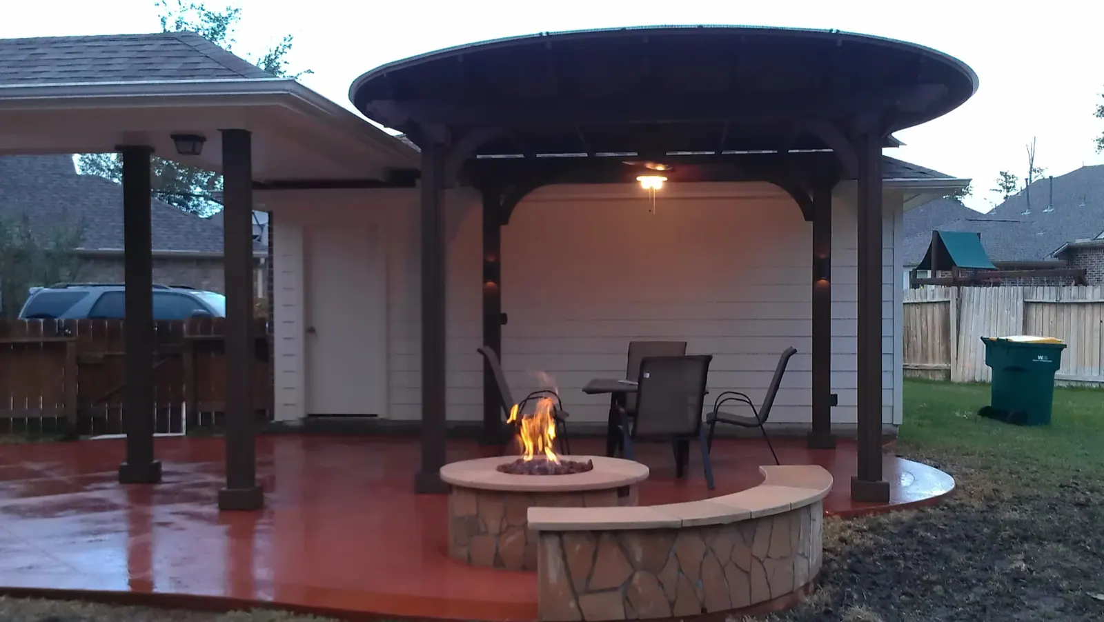 Freestanding round pavilion with stone-clad gas fire pit and curved bench on stamped concrete patio at dusk
