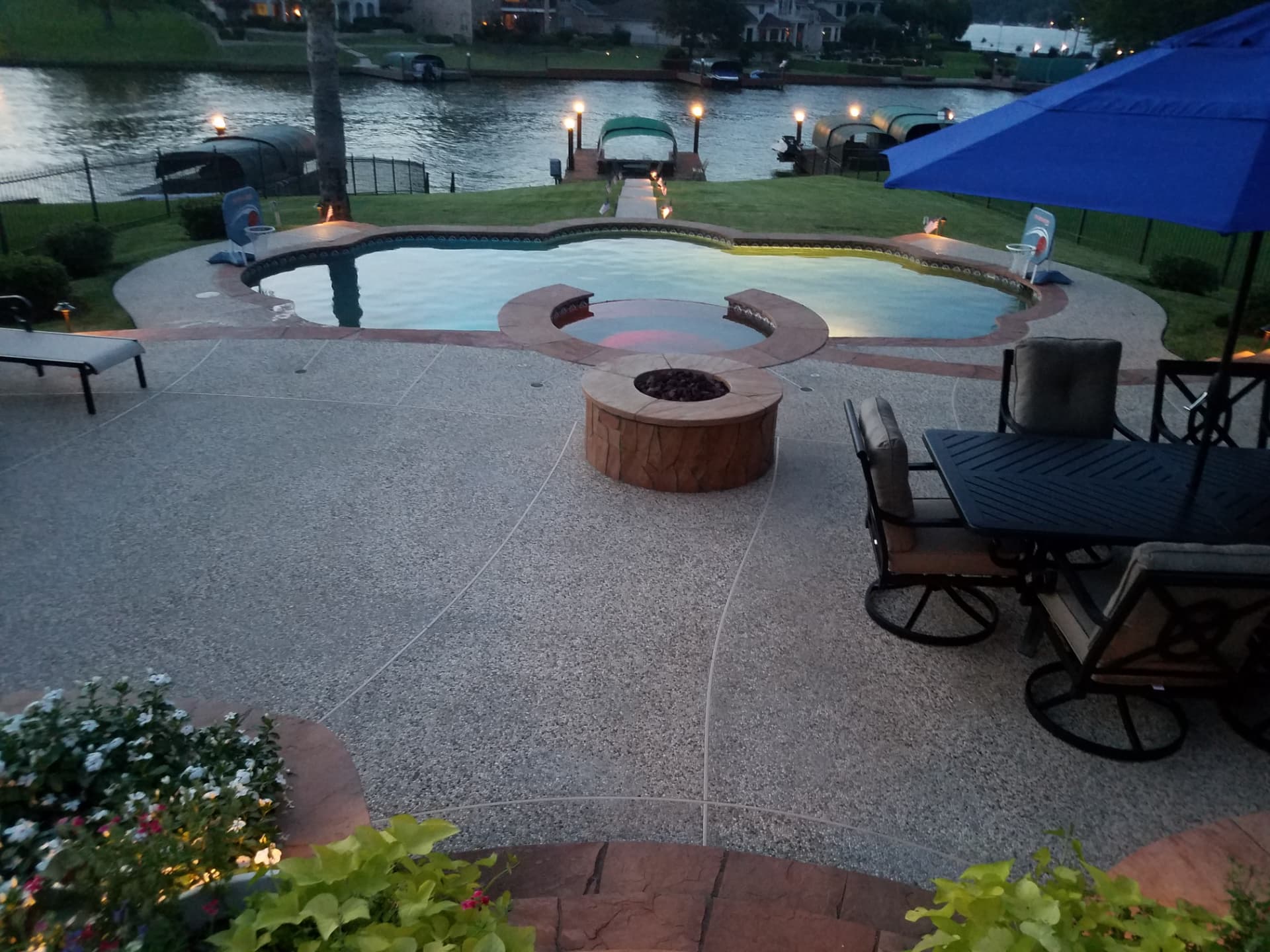 Round flagstone natural gas fire pit on an exposed aggregate patio overlooking a lakeside pool at dusk