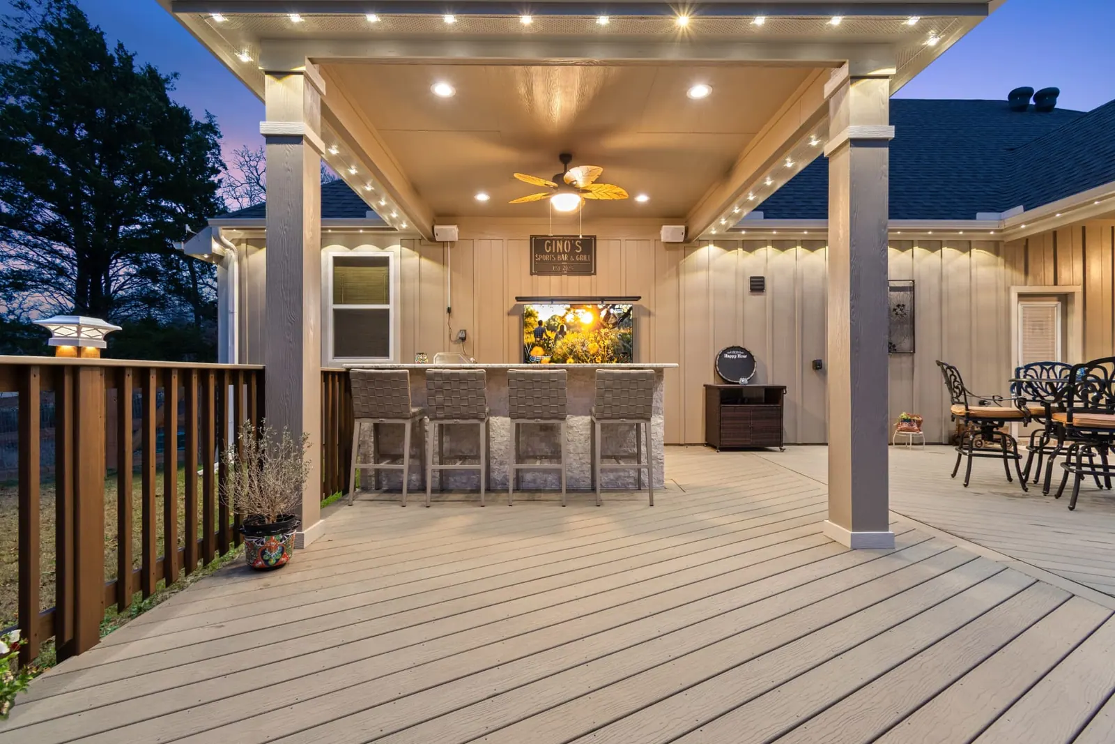 Covered composite deck with stacked-stone bar, sports-bar sign and string lighting at dusk